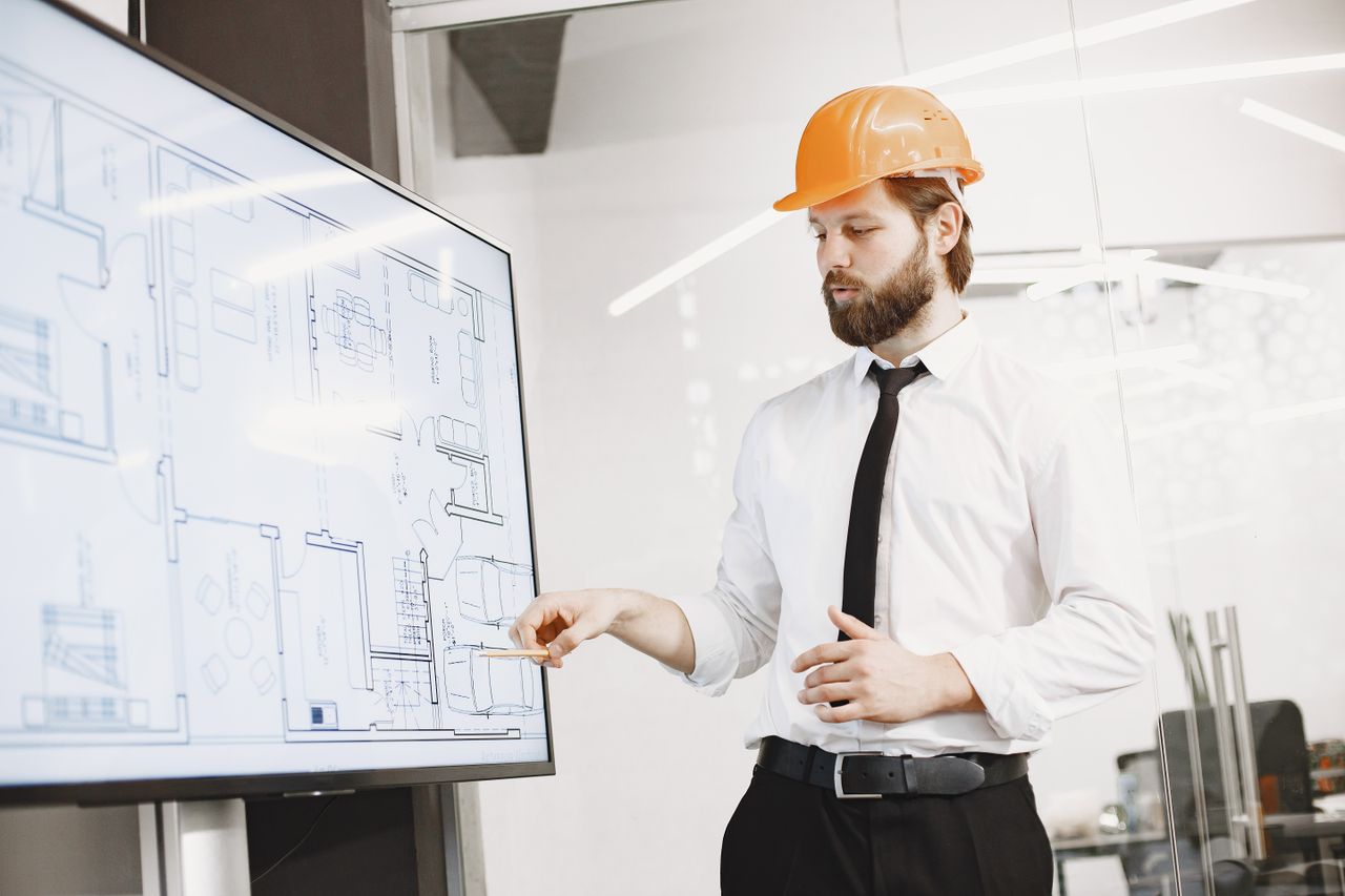 services-04 Architect in hard hat reviewing blueprints on a screen, discussing plans in modern office.